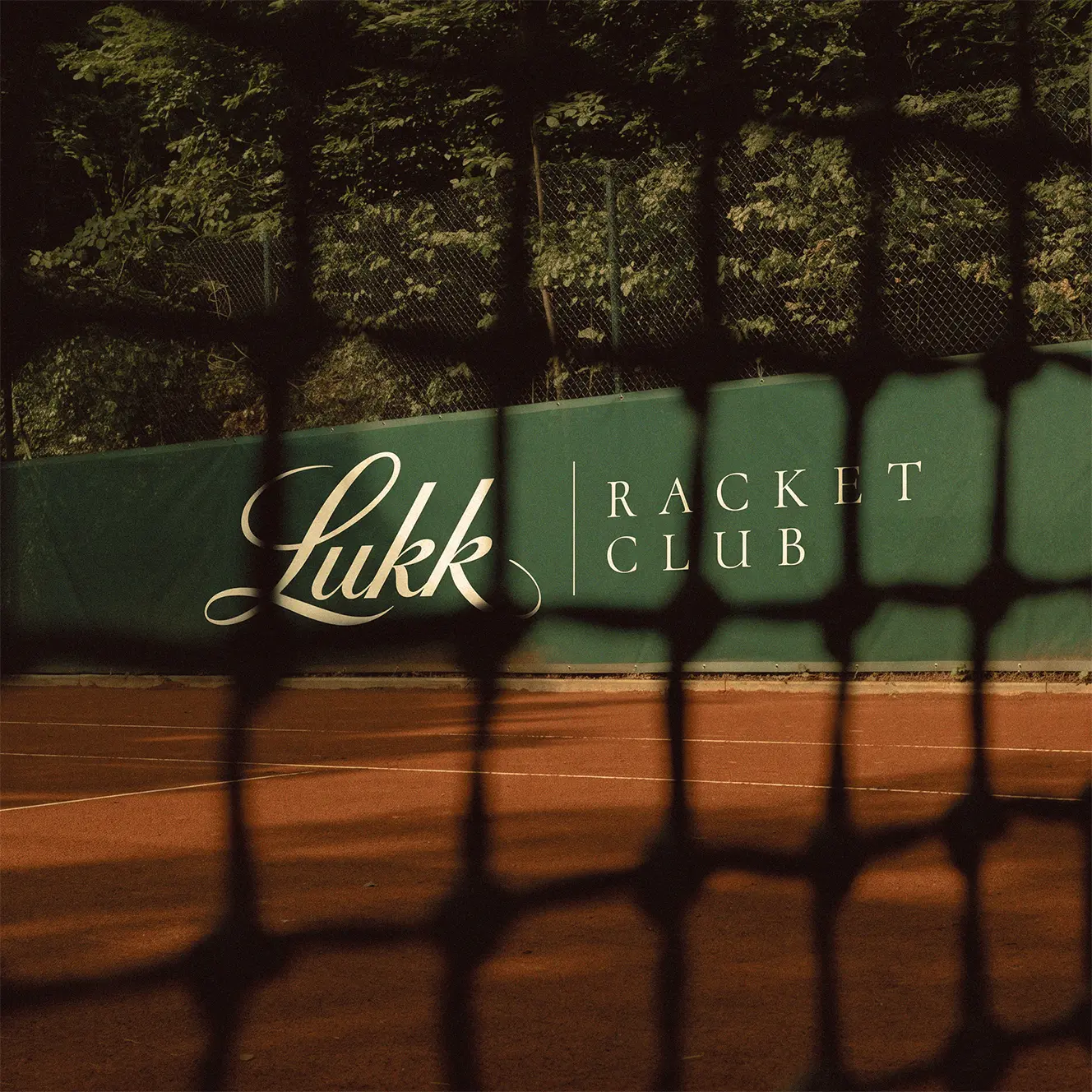 A marca Lukk Racket Club aplicada na lona verde da quadra, com a sombra do alambrado sobre o saibro